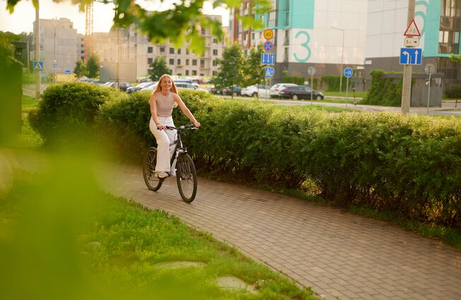 Frau in heller Kleidung fährt auf einem Fahrrad auf einem gepflasterten Weg neben einer Hecke in einem urbanen Gebiet. Mit Link zum Beitrag.