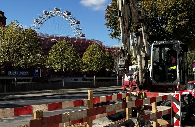 Baustelle mit Bagger, Schubkarre und Absperrung vor Bäumen und einem Riesenrad im Hintergrund