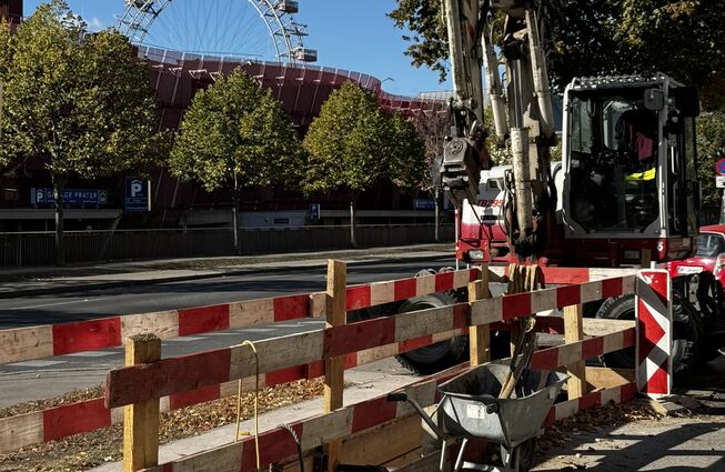 Baustelle mit Bagger, Schubkarre und Absperrung vor Bäumen und einem Riesenrad im Hintergrund
