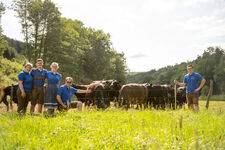 Gruppe von fünf Personen in blauer Arbeitskleidung steht auf einer Wiese neben zwei schwarzen Rindern mit Weidezaun und Wald im Hintergrund