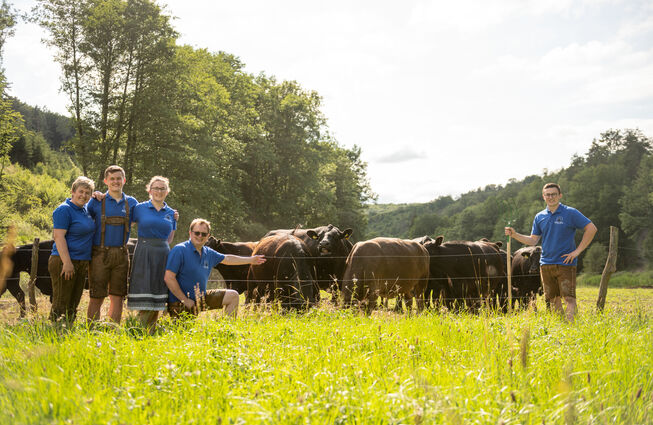 Gruppe von fünf Personen in blauer Arbeitskleidung steht auf einer Wiese neben zwei schwarzen Rindern mit Weidezaun und Wald im Hintergrund