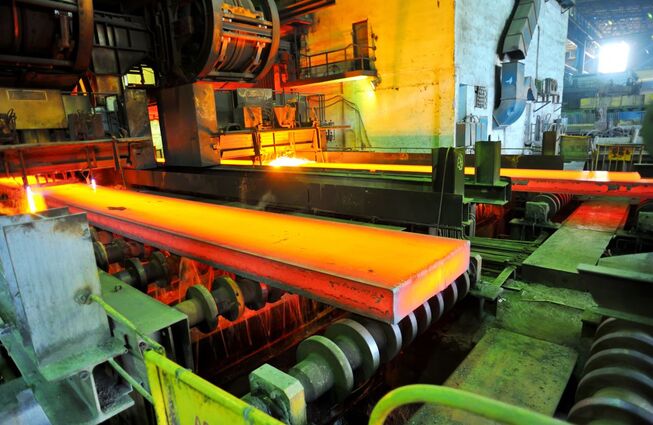 Glühendes Metallstück auf Förderband in einer Industrieanlage mit Maschinen und Rohren im Hintergrund