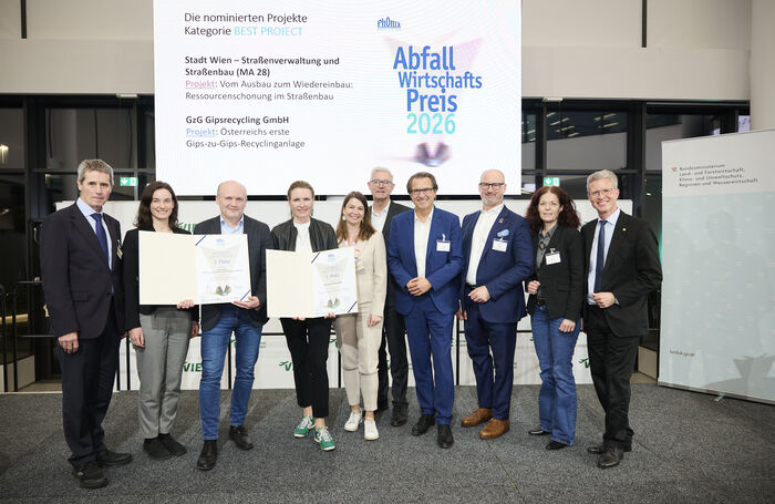 Gruppe von Personen steht vor einer großen Leinwand mit Text zum Abfallwirtschaftspreis 2026 und hält Urkunden in einem Veranstaltungsraum
