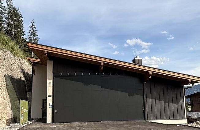 Flaches Gebäude mit schwarzer Garagentür vor bewaldetem Hang und blauem Himmel mit Link zum Beitrag