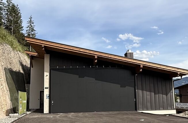 Flaches Gebäude mit schwarzer Garagentür vor bewaldetem Hang und blauem Himmel mit Link zum Beitrag