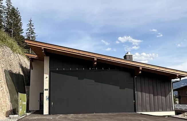 Flaches Gebäude mit schwarzer Garagentür vor bewaldetem Hang und blauem Himmel mit Link zum Beitrag
