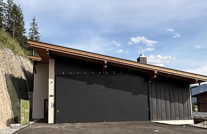 Flaches Gebäude mit schwarzer Garagentür vor bewaldetem Hang und blauem Himmel mit Link zum Beitrag