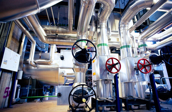 Industrielle Anlage mit mehreren großen, silbernen Rohrleitungen und Ventilen mit schwarzen und roten Handrädern in einer Halle mit Link zum Beitrag.